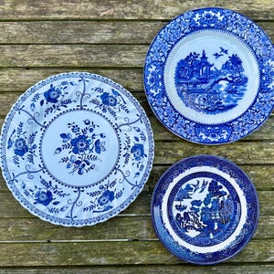 May include: Three blue and white porcelain plates with different floral and landscape designs. The plates are arranged on a wooden surface.