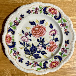 Könnte beinhalten: Weißer Keramikteller mit gewelltem Rand, mit einem Blumendesign in Rosa, Blau und Lila. Der Teller hat einen goldenen Rand und ist mit stilisierten Blumen und Blättern verziert.