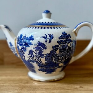 Puede incluir: Una taza de cerámica blanca con un asa negra y el texto "But First, Coffee" en negrita negra. La taza se encuentra sobre un fondo borroso, resaltando su diseño simple.
