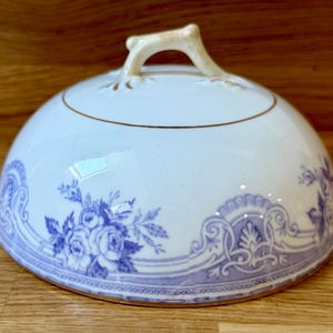 May include: A white ceramic cloche with a blue floral pattern and gold trim. The cloche has a handle on top, likely for serving or display. The floral design features roses and other decorative elements.