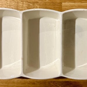 Puede incluir: Una fuente rectangular de cerámica blanquecina con tres compartimentos. Tiene asas verde claro en cada extremo. El plato está diseñado para servir aperitivos o entremeses.