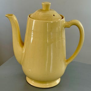 Könnte beinhalten: Eine Vintage-Kaffeekanne aus hellgelber Keramik mit Deckel und Griff. Die Kanne hat einen gebogenen Ausguss und einen abgerundeten Boden. Horizontale Linien umkreisen den Körper. Der Deckel hat einen kleinen Knopf und ein Loch.