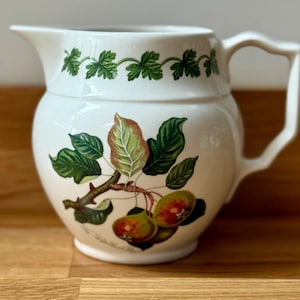 Könnte beinhalten: Weißer Keramikkrug mit einem Feigenzweig-Design. Der Krug hat einen gebogenen Griff und einen Ausguss. Der Feigenzweig hat grüne Blätter und reife Feigen in Grün-, Braun- und Rottönen. Grüner Blättergirlande verziert die Oberseite.