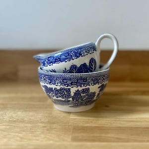 Puede incluir: Un juego de cuenco y crema de cerámica azul y blanca. El cuenco está apilado con la crema, que tiene un asa y un pico. Ambas piezas presentan un patrón tradicional detallado con árboles, edificios y bordes decorativos. El juego está colocado sobre una superficie de madera.