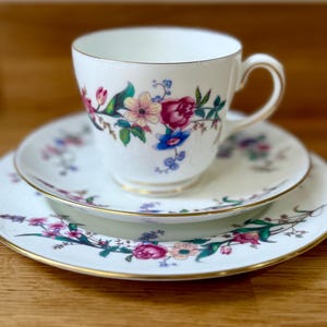 Puede incluir: Juego de té blanco con taza, platillo y plato. Diseño floral con flores rosas, azules y verdes. Bordes dorados. El juego está sobre una superficie de madera.