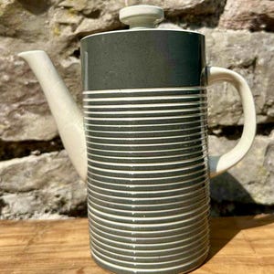 Puede incluir: Una cafetera alta de cerámica con la parte superior gris oscuro y un cuerpo decorado con rayas blancas horizontales. El pico, el asa y la tapa son de color blanquecino. La cafetera está colocada contra una pared de piedra.