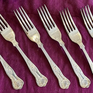 Puede incluir: Seis tenedores de plata con mangos adornados, dispuestos sobre un fondo de tela morado.