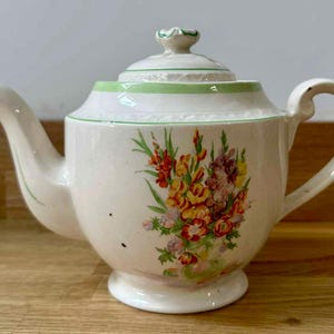 Puede incluir: Tetera de cerámica color crema con un diseño floral en tonos amarillos, rojos y morados. La tetera tiene un pico curvo, un asa redondeada y una tapa con un pequeño pomo. Una banda verde acentúa la parte superior.