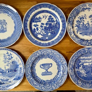 May include: A set of six round ceramic plates featuring various blue and white transferware patterns. The designs include floral motifs, architectural landscapes, a central urn, and traditional willow-style scenes with scalloped edges.