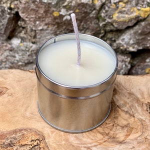 May include: A small, cylindrical candle in a silver metal container. The candle has a white wax and a single, centered wick. The container sits on a wooden surface, with a stone wall in the background.