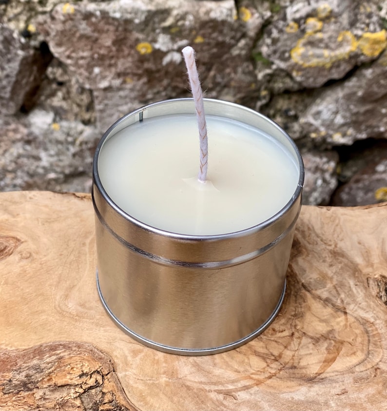May include: A small, cylindrical candle in a silver metal container. The candle has a white wax and a single, centered wick. The container sits on a wooden surface, with a stone wall in the background.