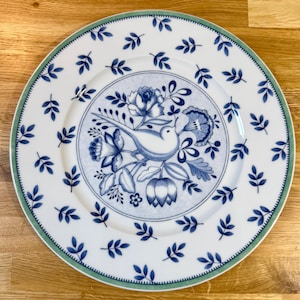 May include: White ceramic plate with a blue and white floral and bird design. The plate features a central motif of a bird perched among flowers, surrounded by a border of blue leaves and a green rim. The plate is on a wooden surface.