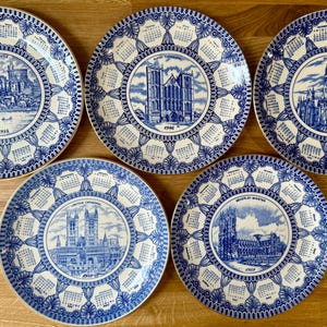 May include: Five vintage decorative plates with blue and white designs. Each plate features a central image of a cathedral or castle, surrounded by a calendar-style border. The plates are arranged on a wooden surface.