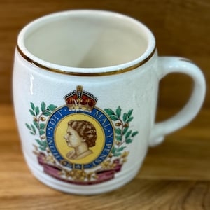 Op de afbeelding: Crèmekleurige keramische mok met een gouden rand en oor. De mok heeft een wapen met een portret, kroon en de woorden "HONI SOIT QUI MAL Y PENSE". Een decoratief item voor in de keuken of als verzamelobject.