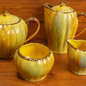 Pode incluir: Um conjunto de chá vintage com bule, cremeira, açucareiro e jarro. O conjunto é amarelo com acabamentos e alças douradas. O bule tem uma tampa com um botão dourado. O conjunto está sobre uma superfície de madeira.