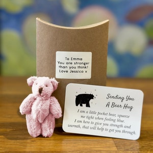 Può includere: Un orsacchiotto rosa con un biglietto che dice "Sending You A Bear Hug" e include il testo "I am a little pocket bear, squeeze me tight when feeling blue. I am here to give you strength and warmth, that will help to get you through."