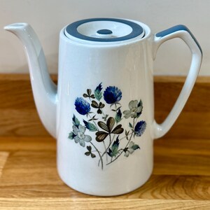 Peut inclure: Théière en céramique blanche avec des détails bleus sur le bord, la poignée et le couvercle. Motif floral avec des fleurs et des feuilles bleues, grises et marron. Le bec verseur est incurvé et la poignée est angulaire.