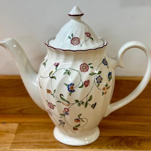 Könnte beinhalten: Cremefarbene Keramik-Teekanne mit Deckel, verziert mit einem Blumenmuster in Rosa, Blau und Orange. Die Teekanne hat einen gebogenen Griff und Ausguss sowie einen roten Rand am Deckel.