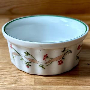 Pode incluir: Uma ramequim de cerâmica branca com borda ondulada e borda verde. O exterior é decorado com uma fita rosa e pequenos detalhes florais vermelhos. A ramequim é projetada para assar e servir porções individuais.