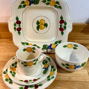 May include: A cream-colored ceramic dish set with a square serving platter, a cup and saucer, a small plate, and a bowl. Each piece is decorated with a pattern of yellow oranges, green leaves, and purple grapes. The set has a vintage aesthetic.