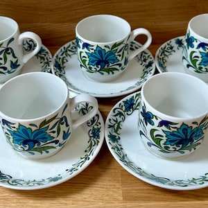 Op de afbeelding: Set van vijf witte keramische kopjes en schotels. Elk kopje en schotel heeft een blauw en groen bloemmotief. De kopjes hebben een oor en de schotels zijn rond. De set is geplaatst op een houten oppervlak.