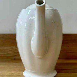 May include: Cream-colored ceramic teapot with a curved spout and handle. The teapot has a rounded body and a lid. The teapot is sitting on a wooden surface.
