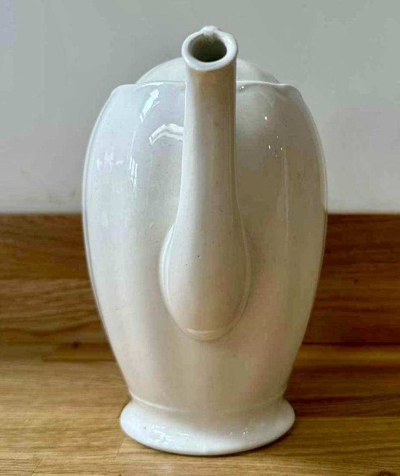 May include: Cream-colored ceramic teapot with a curved spout and handle. The teapot has a rounded body and a lid. The teapot is sitting on a wooden surface.