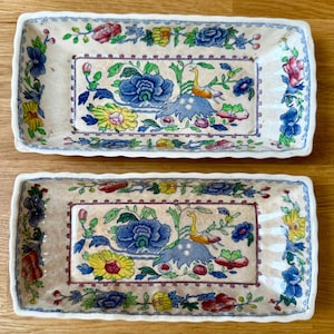 Op de afbeelding: Twee rechthoekige keramische dienbladen met een crèmekleurige basis en een kleurrijk bloemenpatroon. Het ontwerp bevat blauwe, gele, roze en groene bloemen, samen met een centraal vogelmotief, omlijst door een blauw-witte decoratieve rand.