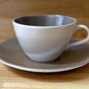 Puede incluir: Un juego de taza y platillo de cerámica de tonos neutros. La taza tiene una forma redondeada con un asa y un interior más oscuro. El platillo es de color a juego con un borde ligeramente texturizado. El juego está diseñado para servir bebidas calientes.