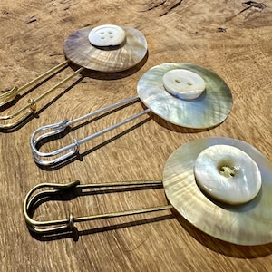 Puede incluir: Tres imperdibles decorativos con botones de aspecto perlado. Cada imperdible tiene un acabado metálico de color diferente: dorado, plateado y bronce.