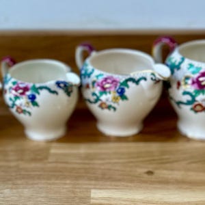 May include: Four cream-coloured ceramic cream jugs with floral designs. Each jug has a handle with a burgundy accent and a spout. The floral pattern includes pink, blue, and yellow flowers with green leaves. The jugs are arranged in a row.