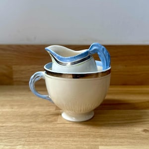 May include: A cream-colored teacup and creamer set. The cup has a gold rim and a light blue handle. The creamer, nested inside, has a blue handle and a blue and silver rim. The set is on a wooden surface.
