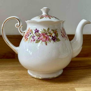 Puede incluir: Tetera de porcelana blanca con diseño floral en rosa, morado y amarillo. La tetera tiene un asa, un pico y una base con ribetes dorados. La tapa también está decorada con flores y tiene un pequeño pomo.