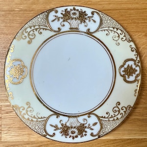 Könnte beinhalten: Ein runder, cremefarbener Teller mit goldenem Blumen- und Scroll-Design. Der Teller hat einen zentralen weißen Kreis, einen goldenen Rand und goldene Blumenkörbe und Scroll-Akzente. Der Teller ist wahrscheinlich aus Porzellan oder Keramik.