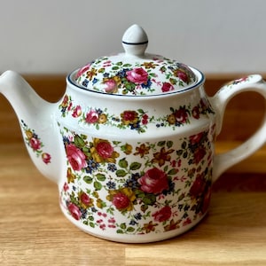 May include: A white ceramic teapot with a lid, adorned with a floral design featuring red roses, yellow blooms, and green foliage. The teapot has a curved spout and handle, with a blue trim around the lid.