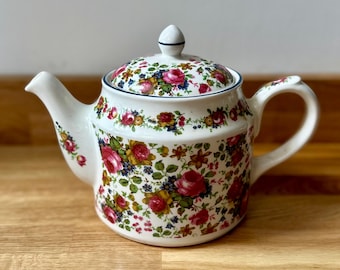May include: A white ceramic teapot with a lid, decorated with a floral pattern of red roses, yellow flowers, and green leaves. The teapot has a curved spout and handle, and a blue trim around the lid.
