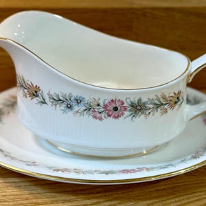 May include: A white porcelain gravy boat with a matching saucer. The gravy boat has a curved spout and a handle. Both pieces are decorated with a floral pattern in pink, blue, and gold. The rims are trimmed with gold.