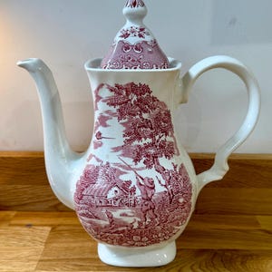 May include: A white ceramic coffee pot with a lid, decorated with a red and white pastoral scene. The pot has a curved spout and handle, and the lid is topped with a decorative finial. The design includes a hunter, trees, and a building.