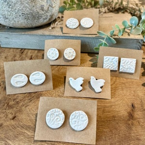 Op de afbeelding: Assortiment van witte keramische oorbellen op bruin karton. De oorbellen hebben verschillende ontwerpen, waaronder bloemen, vogels en geometrische patronen.