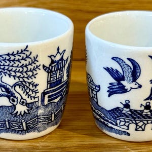 May include: Two white ceramic cups with a blue Willow pattern design. One cup features a weeping willow tree and a pagoda, while the other depicts birds in flight and a bridge. The cups are cylindrical and have a smooth finish.