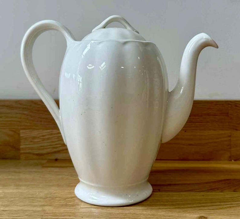 May include: A white ceramic teapot with a curved spout and handle. The teapot has a lid with a small handle and a ribbed design on the body. The teapot is sitting on a wooden surface.