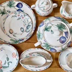 May include: A ceramic dinnerware set featuring a white base with a colorful botanical pattern of figs, green leaves, and blossoms. The collection includes a large platter, plates, a lidded tureen, a creamer, a sugar bowl, a gravy boat, and a teacup.