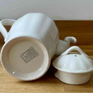 May include: A white ceramic teapot with a matching lid. The teapot has a curved handle and spout. The lid has a small handle. The bottom of the teapot has a stamp that reads "Cream Petal" and "England".