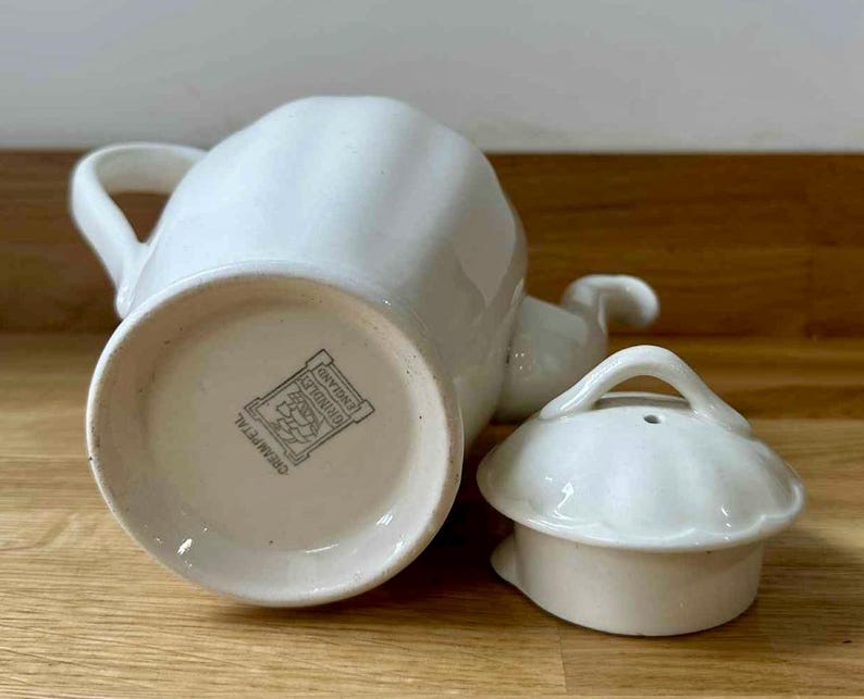 May include: A white ceramic teapot with a matching lid. The teapot has a curved handle and spout. The lid has a small handle. The bottom of the teapot has a stamp that reads "Cream Petal" and "England".