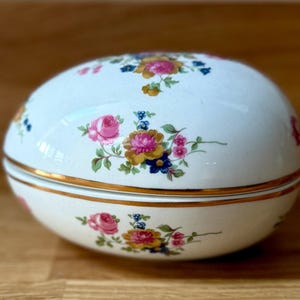 Puede incluir: Una caja de baratijas de cerámica blanca en forma de huevo con tapa, adornada con patrones florales en rosa, amarillo y azul. La caja tiene un borde dorado y está sobre una superficie de madera.