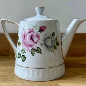 Könnte beinhalten: Weiße Keramik-Teekanne mit Deckel, mit rosa und grauen Rosenblüten-Designs und goldenen Verzierungen. Die Teekanne hat einen gebogenen Griff und einen Ausguss. Der Sockel hat ein dekoratives Muster.