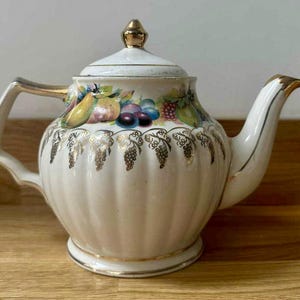 Puede incluir: Tetera de cerámica color crema con tapa y detalles dorados. La tetera presenta un diseño de frutas con uvas, peras y ciruelas. El asa y el pico también están adornados con oro. La tetera está sobre una superficie de madera.