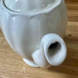 May include: A white ceramic teapot with a lid, spout, and handle. The teapot is on a wooden surface. The teapot has a smooth, glossy finish and a classic design. The spout is curved and the handle is rounded.
