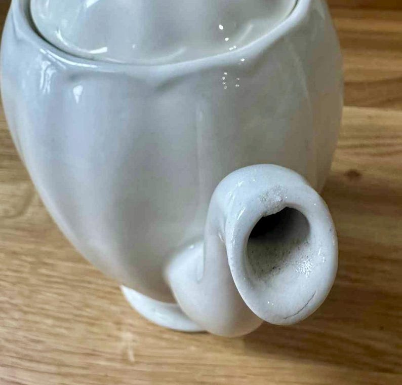 May include: A white ceramic teapot with a lid, spout, and handle. The teapot is on a wooden surface. The teapot has a smooth, glossy finish and a classic design. The spout is curved and the handle is rounded.