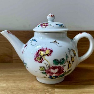 Puede incluir: Tetera de cerámica blanca con tapa, decorada con coloridos diseños florales y de libélulas. La tetera tiene un pico curvo y un asa redondeada. El patrón floral incluye flores rojas, amarillas y verdes.